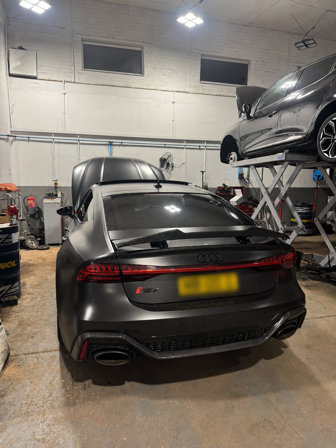 Dark performance car in the garage with its bonnet open and another vehicle on a lift